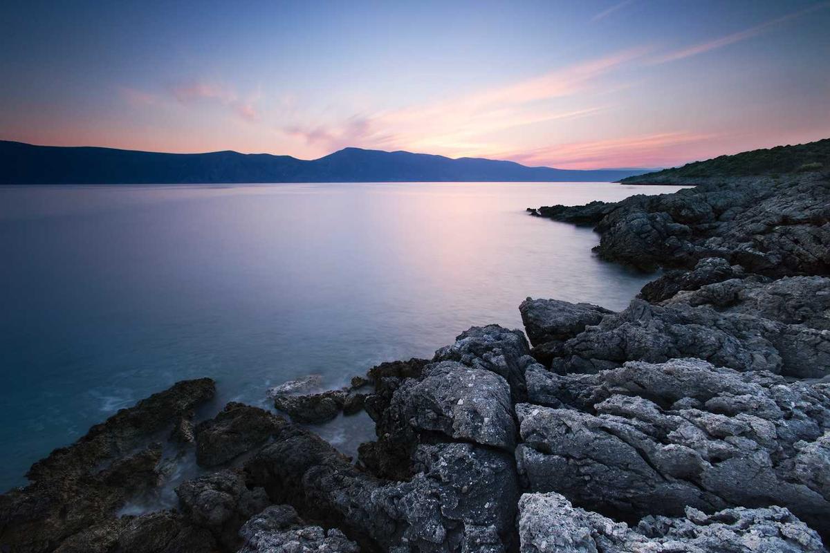Rocky coastline meets calm water, mountains in distance.