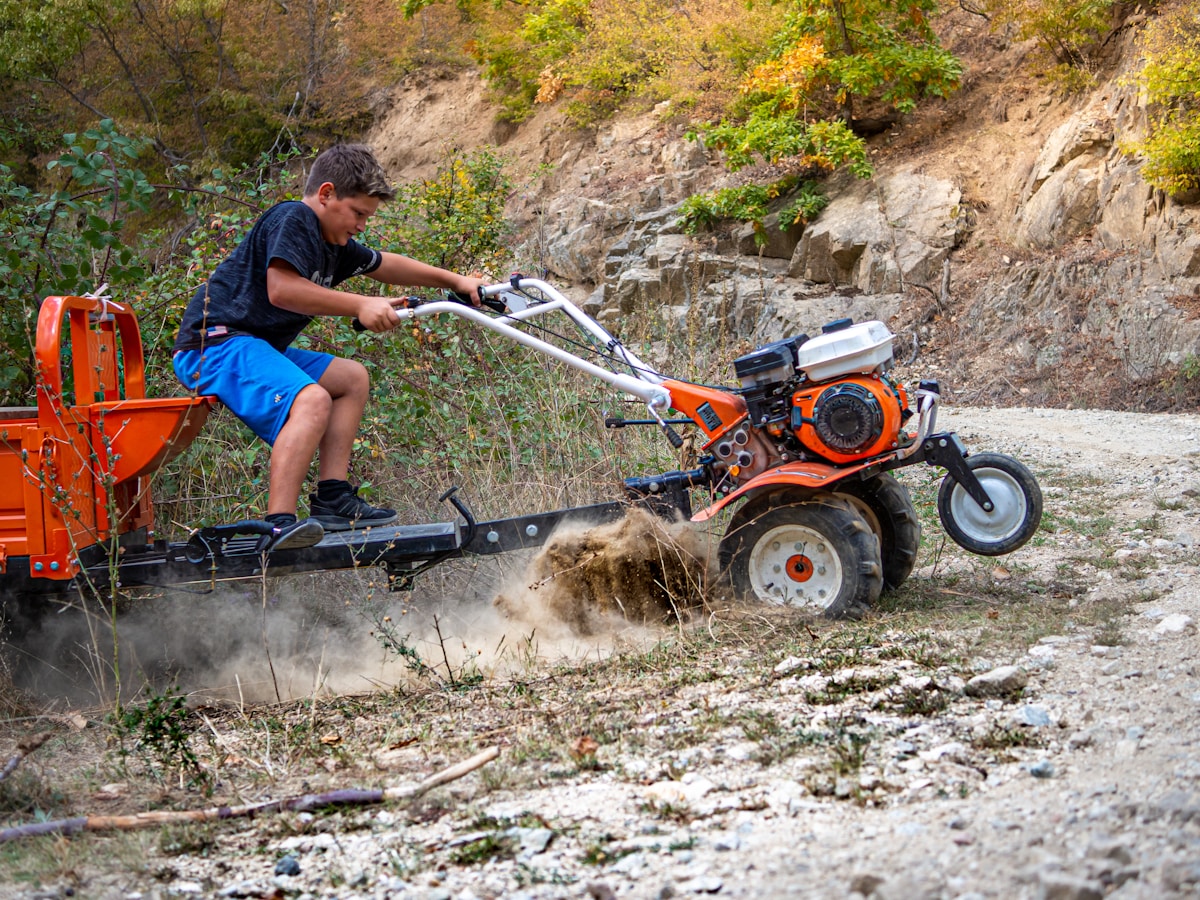 Un homme conduit un tracteur orange pour déblayer un chemin de terre dans un paysage forestier.