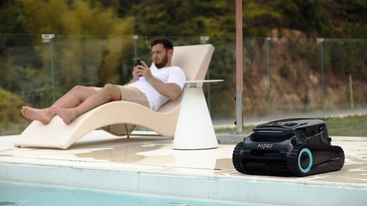 Homme assis sur une chaise longue, utilisant un smartphone, près d'une piscine et d'un robot de piscine.