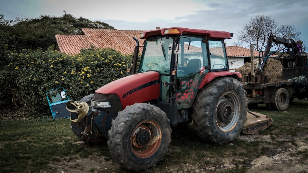 Tracteur agricole rouge garé dans un champ avec des bâtiments en arrière-plan.