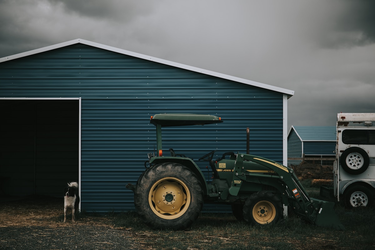 Tracteur vert John Deere garé devant un hangar bleu sous un ciel orageux.