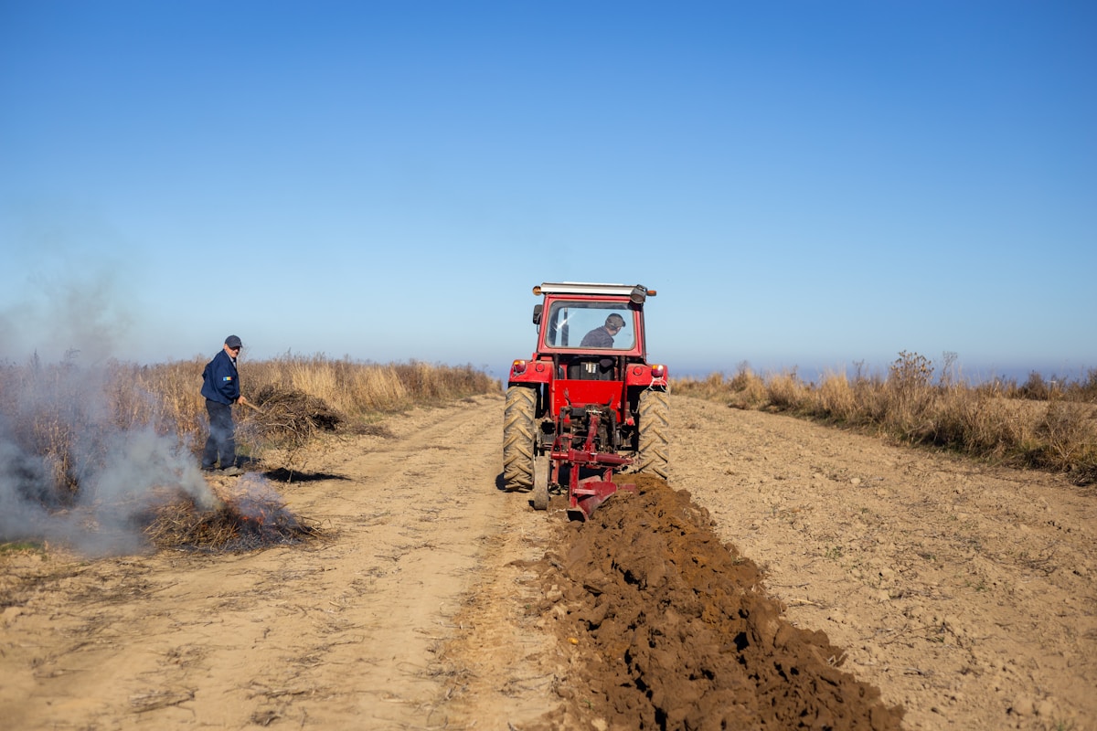 Tracteur rouge labourant un champ avec un homme debout à proximité.