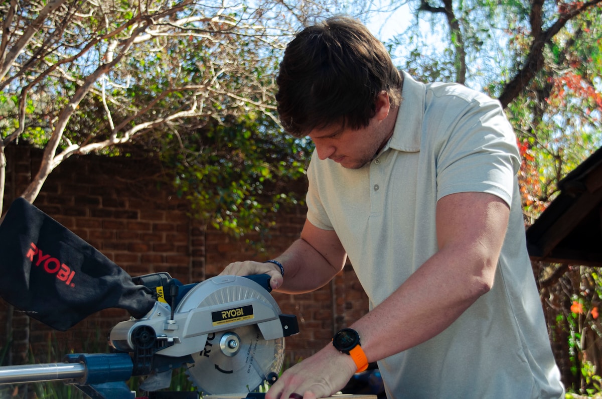 Homme utilisant une scie circulaire Ryobi dans un jardin verdoyant.